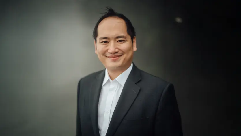 A smiling man in a suit stands against a dark background, exuding confidence and professionalism.