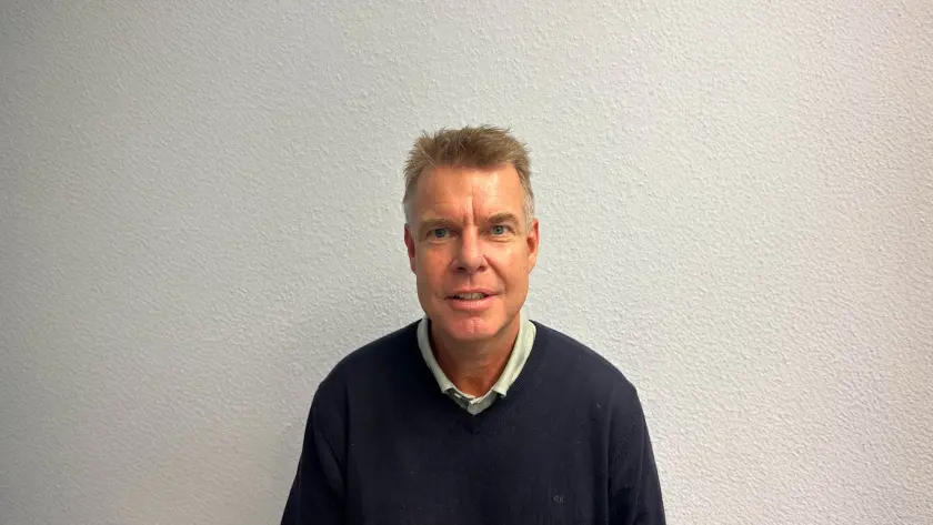 A man with short hair, wearing a dark sweater over a collared shirt, smiles against a light textured wall.