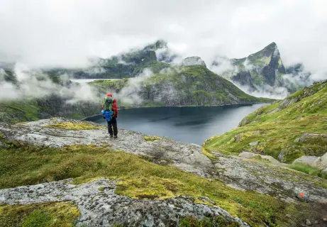 A hiker in a red jacket stands on rocky terrain overlooking a serene lake surrounded by lush green mountains and misty clouds.