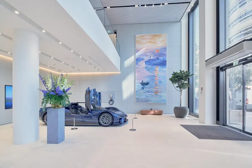 A luxury car with gullwing doors is displayed in a modern lobby featuring a large artwork, potted plants, and bright natural light.