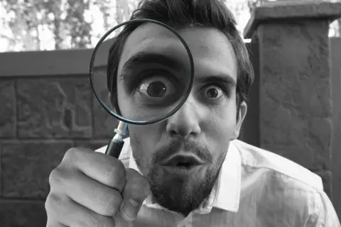 A man is holding a magnifying glass to his eye, making a surprised expression, with a blurred outdoor background.