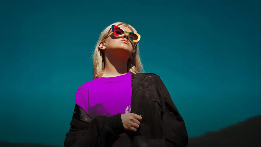 A woman with blonde hair wears colorful sunglasses and a purple shirt, standing confidently against a dark blue background.