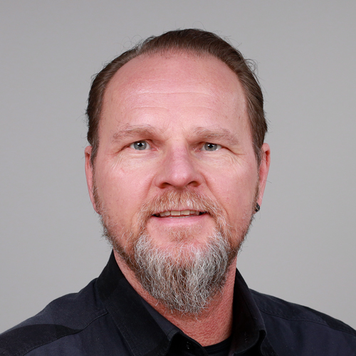 A middle-aged man with a beard and short hair smiles at the camera, wearing a black shirt against a neutral background.