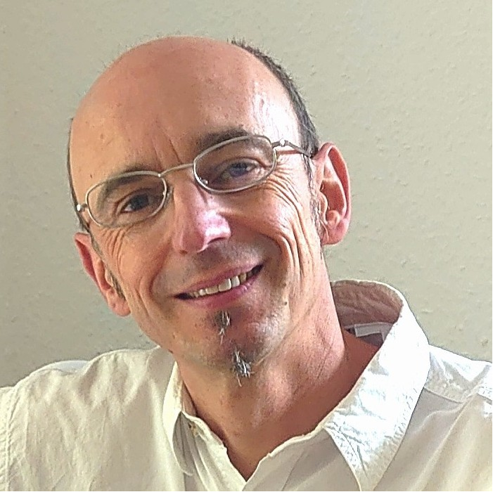 A smiling man with glasses and a goatee, wearing a white shirt, poses against a light-colored wall.