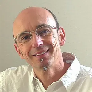 A smiling man with glasses and a goatee, wearing a white shirt, poses against a light-colored wall.