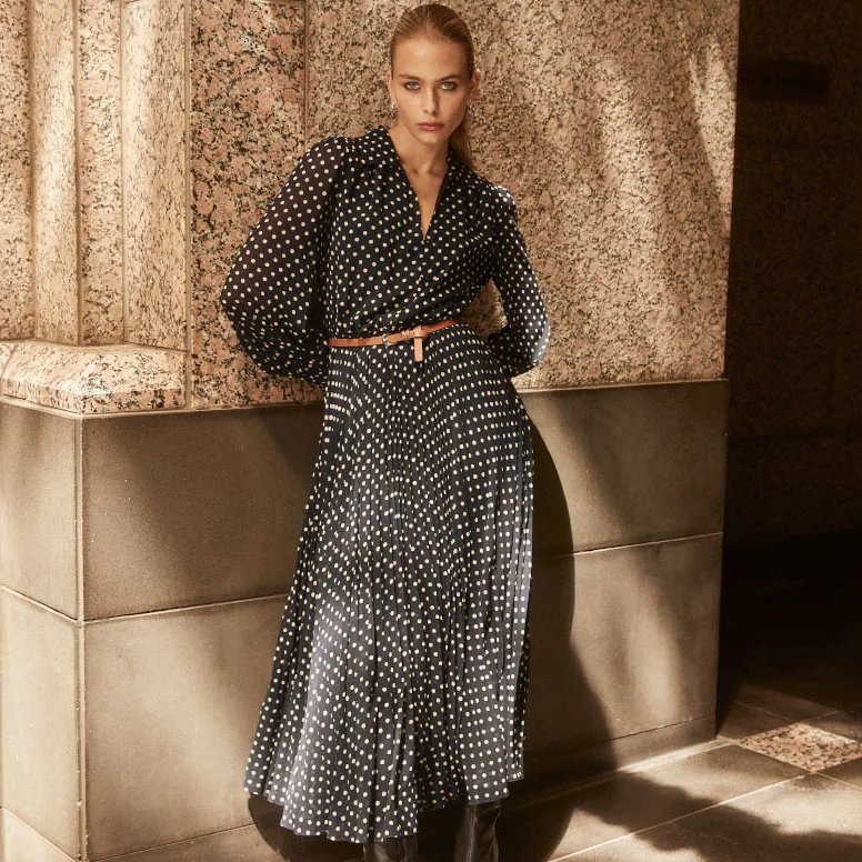 A model poses against a textured wall, wearing a polka dot dress with a belt, showcasing a stylish and elegant look.