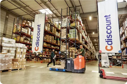 A warehouse with tall shelves stacked with boxes; two workers maneuvering equipment among the aisles. A 'Cdiscount' banner is visible.