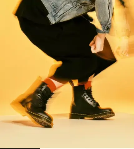 A person in a denim jacket and black skirt is mid-step, wearing black combat boots with yellow soles, against a bright yellow background.