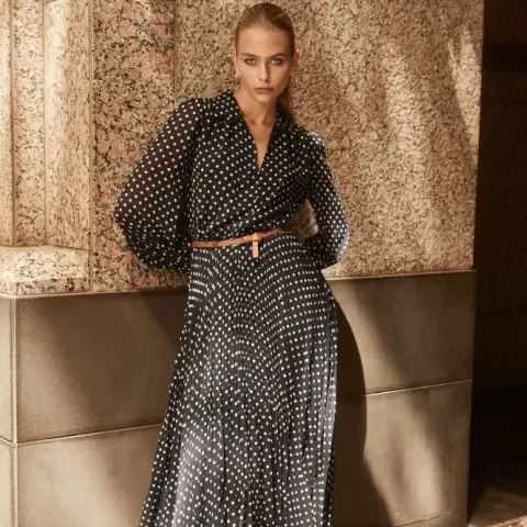 A model poses against a textured wall, wearing a polka dot dress with a belt, showcasing a stylish and elegant look.