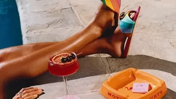 A pair of colorful sandals rests beside a pink cocktail and an orange ashtray labeled 'MOTEL KURT'S,' with a hand resting on the edge.