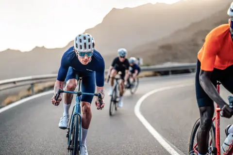 A group of cyclists races on a winding road, with one rider in a blue outfit leading the pack against a mountainous backdrop.