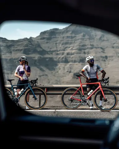 Two cyclists, a woman in a purple and black outfit and a man in a gray and orange outfit, rest by their bikes against a mountainous backdrop.
