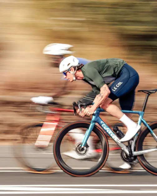 Two cyclists race on a road, one in a green jersey and blue bike, the other blurred in red, showcasing speed and motion.