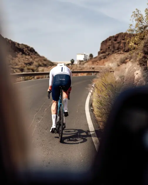 A cyclist in a blue and white outfit rides on a winding road surrounded by rocky terrain and sparse vegetation.