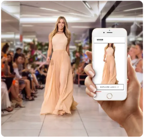 A model walks the runway in a flowing peach gown while a hand holds a smartphone displaying the outfit for viewers.