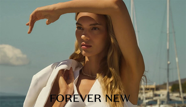 A woman with long hair poses outdoors, wearing a white outfit, with a marina and boats in the background. The text 'FOREVER NEW' is displayed prominently.