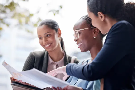 Three women are engaged in discussion, reviewing documents outdoors, smiling and pointing at the papers.