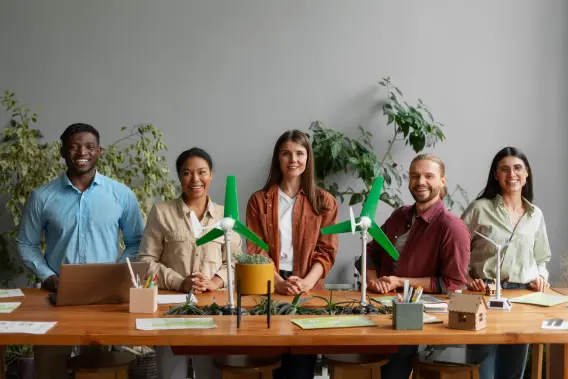 A diverse group of five people stands behind a table with wind turbine models, plants, and a laptop, smiling in a bright, green space.