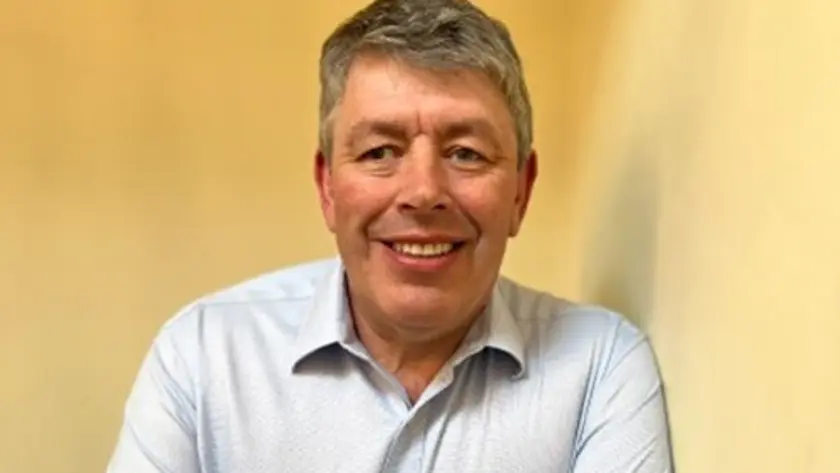 A smiling man with short gray hair stands with arms crossed, wearing a light blue button-up shirt against a neutral background.