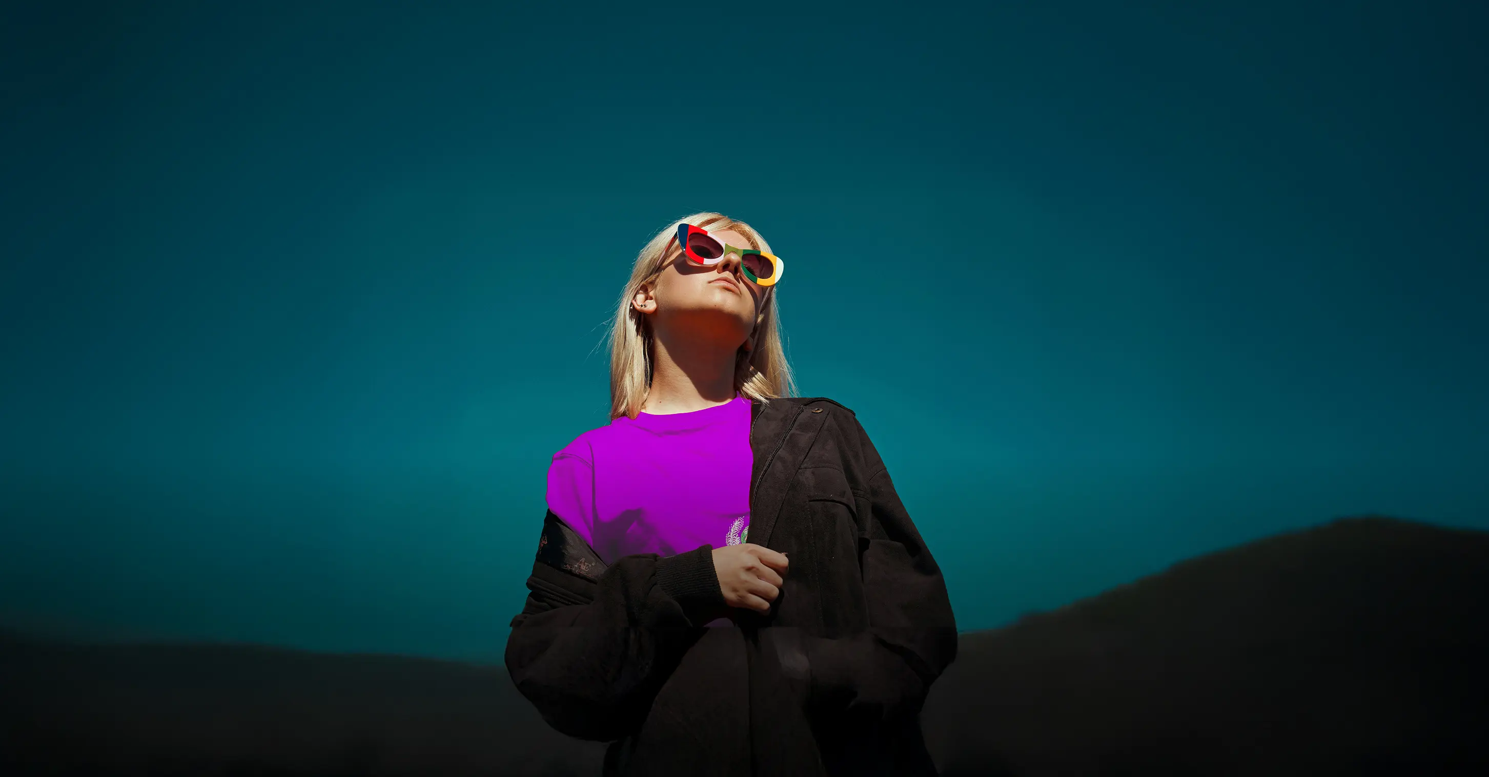 A person with blonde hair wears colorful sunglasses and a purple shirt, standing against a dark blue sky and distant hills.