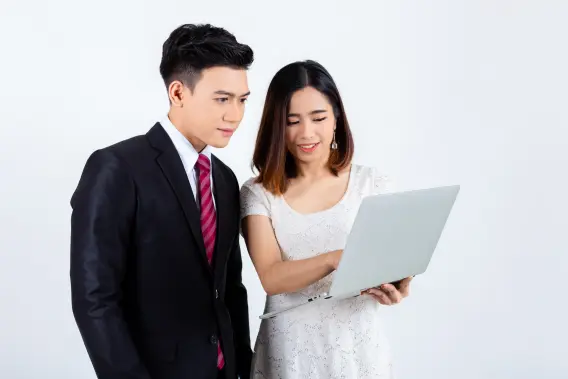 A man in a suit and a woman in a white dress are looking at a laptop, discussing something with smiles on their faces.