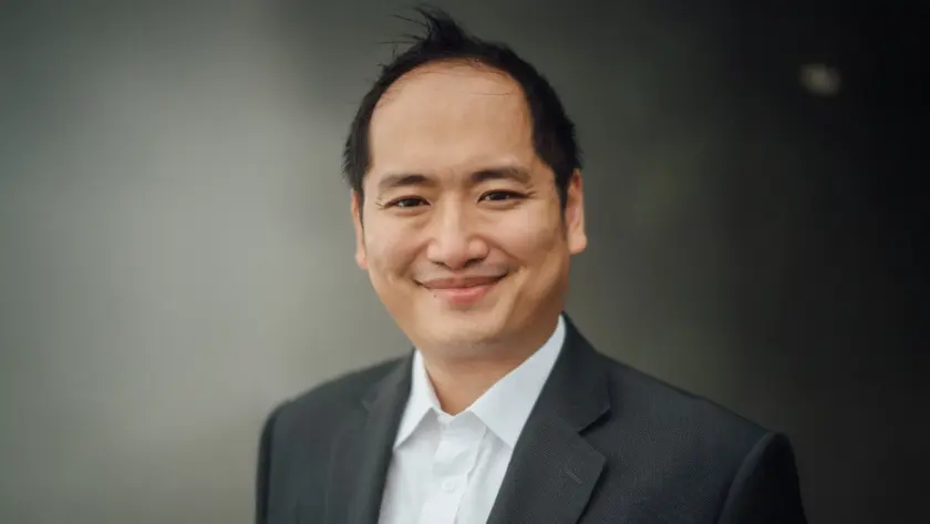 A smiling man in a suit stands against a dark background, exuding confidence and professionalism.