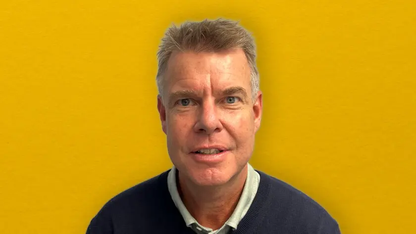 A man with short hair smiles, wearing a dark sweater, against a bright yellow background.