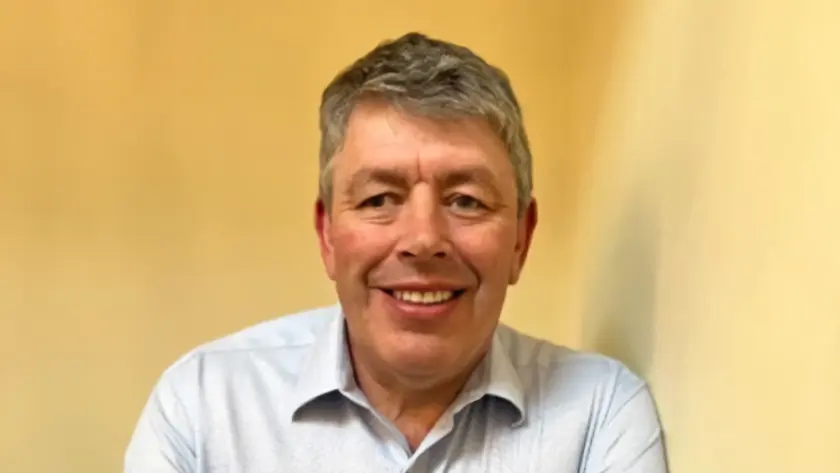 A smiling man with short gray hair stands against a light-colored wall, wearing a light blue shirt and crossing his arms.