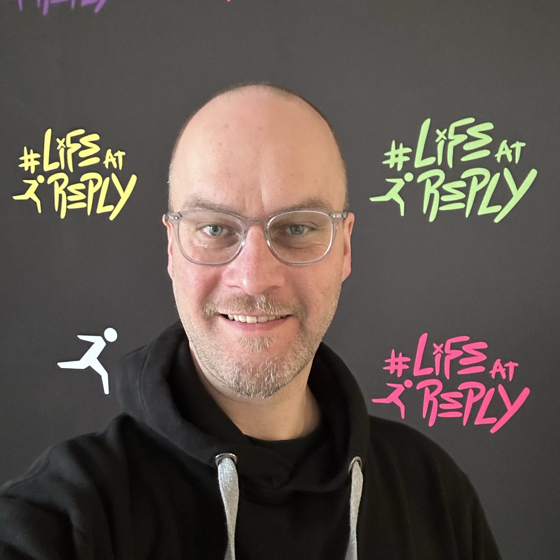 A smiling man with glasses poses in front of a black backdrop featuring colorful text and a running figure, promoting 'Life at Reply.'